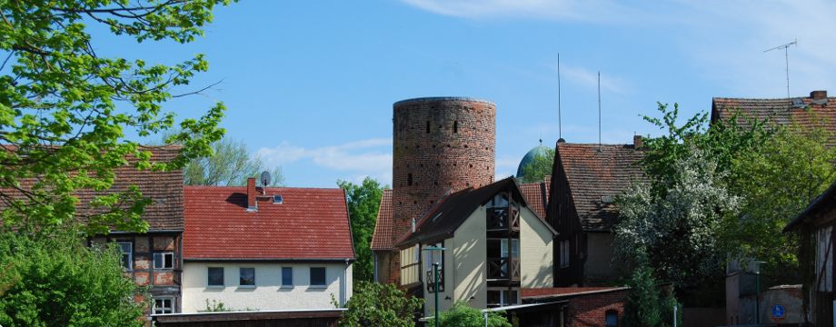 Urlaubsregion Höhbeck Elbe - Titelbild: Häuseransicht mit "Stumpfer Turm", einst Teil der Stadtmauer, komplett aus Backstein, zwischen den Häusern stehen Bäume, im Hintergrund sieht man die Spitze des Burgturmes.